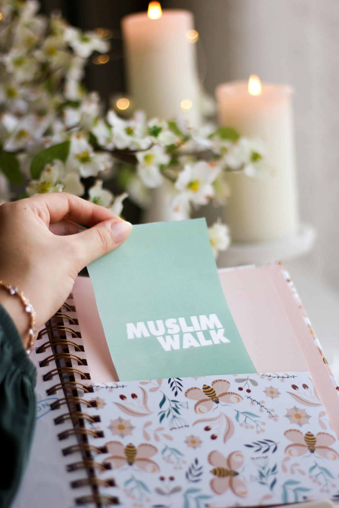 A hand holds a 'Muslim Walk' card above a floral-patterned notebook with candles and flowers in the background.